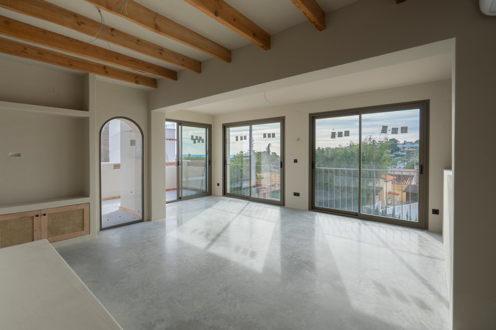 Bright living area featuring exposed wooden beams, polished concrete floors, and large sliding doors with sea views. Built-in shelving with natural clay finishes adds texture.