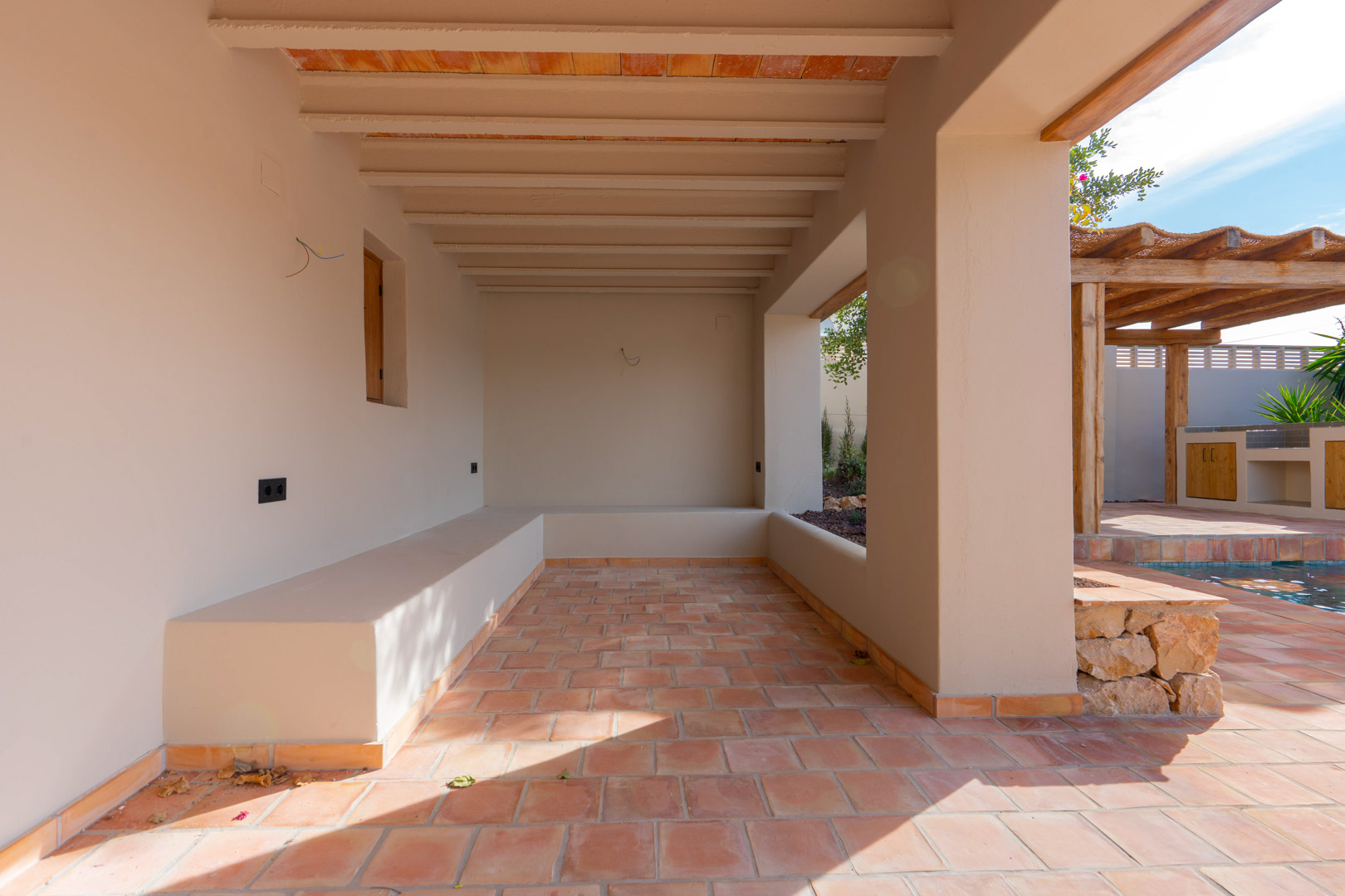 Covered porch featuring terracotta flooring, exposed ceiling beams, and integrated masonry seating overlooking the pool and pergola area.