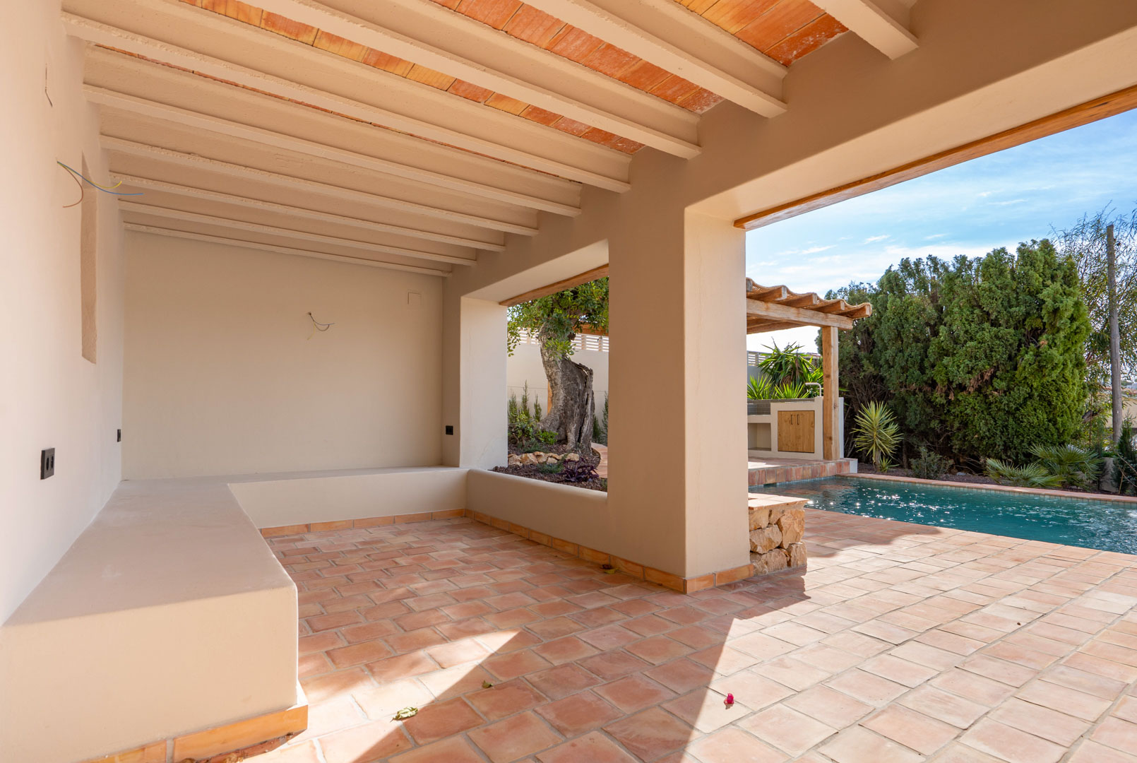 Covered terrace featuring terracotta flooring, exposed ceiling beams, and a built-in masonry bench overlooking the pool and outdoor pergola.