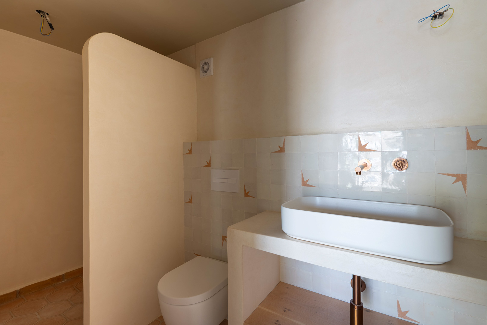 Designer bathroom featuring a vessel sink on a masonry vanity, copper wall-mounted fixtures, and clay plaster walls. Includes a wall-hung toilet and terracotta flooring.
