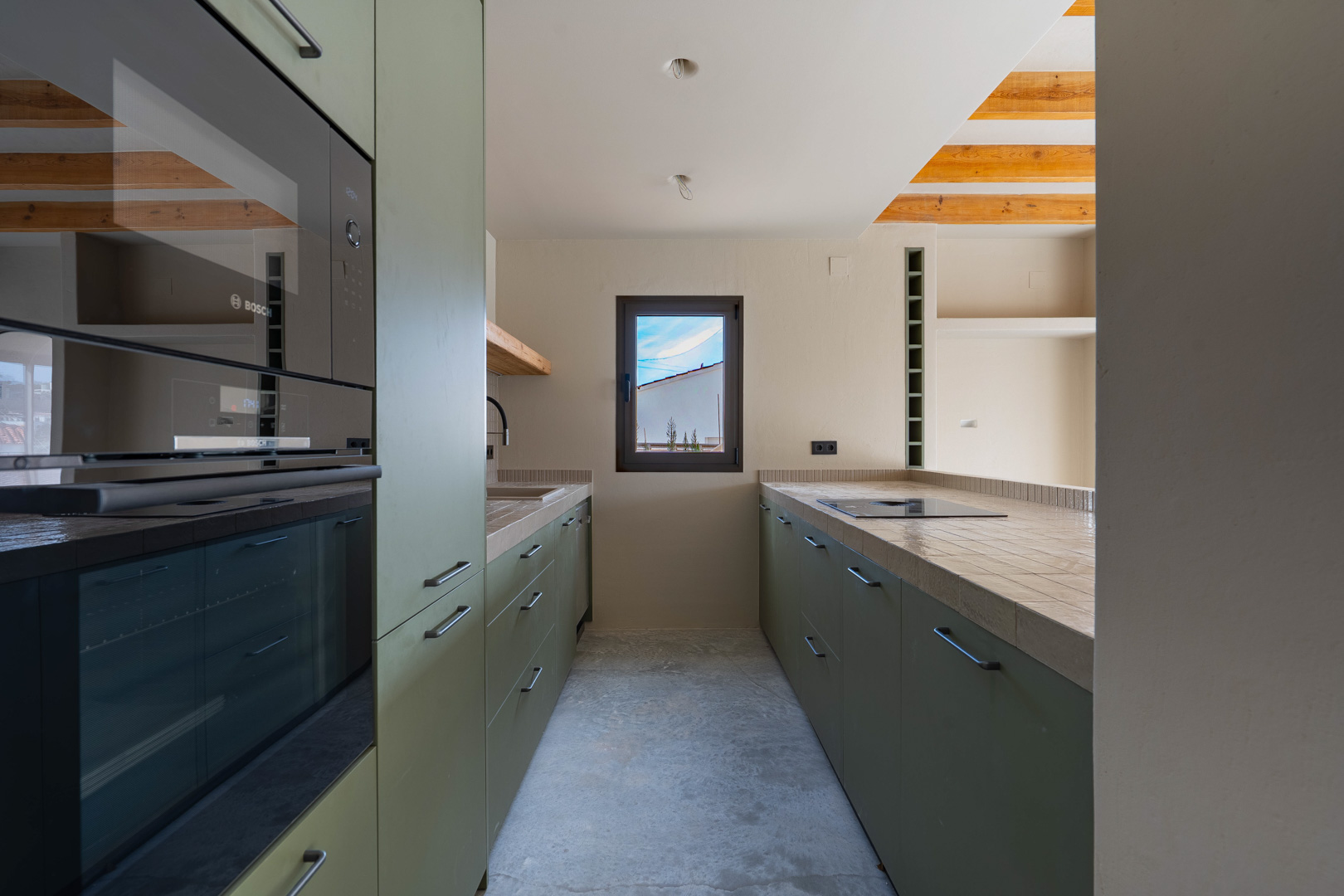 Designer kitchen with olive green cabinetry, integrated Bosch appliances, and porcelain tiled countertops. Features exposed wooden beams and polished concrete flooring.