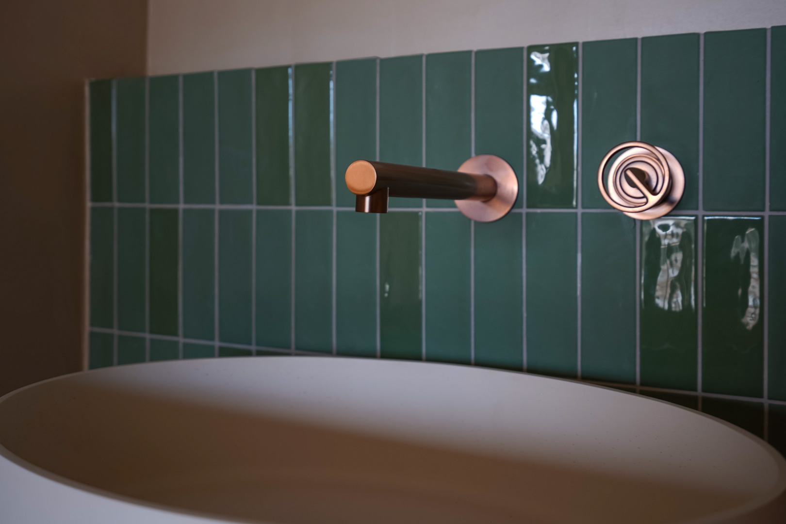 Detail of a bathroom featuring a wall-mounted copper faucet and controls set against glossy green vertical tiles and a matte white vessel sink.
