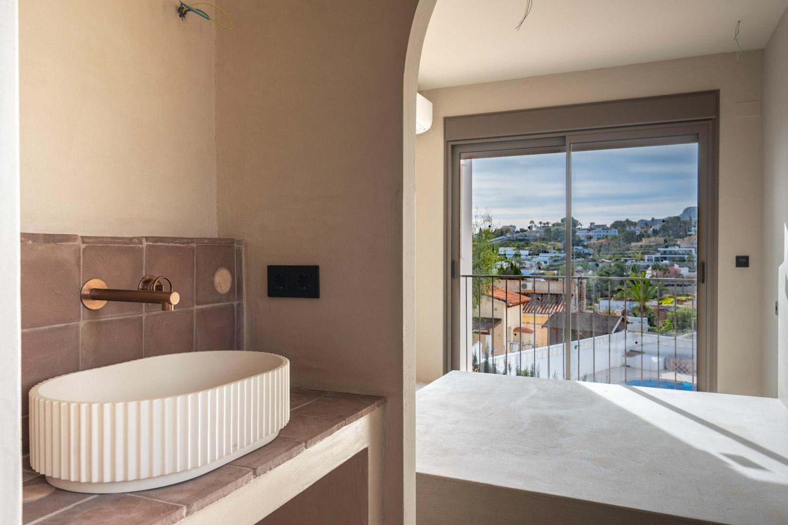 Ensuite bathroom featuring a fluted vessel sink and copper fixtures, viewed through an archway leading to the bedroom with balcony and landscape views.