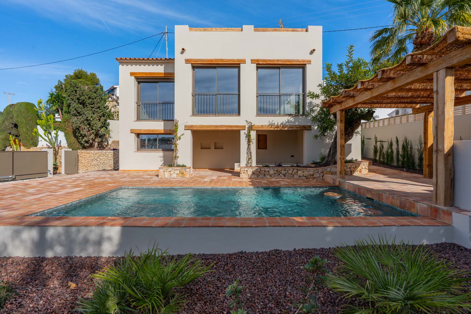 Exterior view featuring a rectangular swimming pool with terracotta tiling, a rustic wooden pergola, and a white facade accented by natural wood lintels.