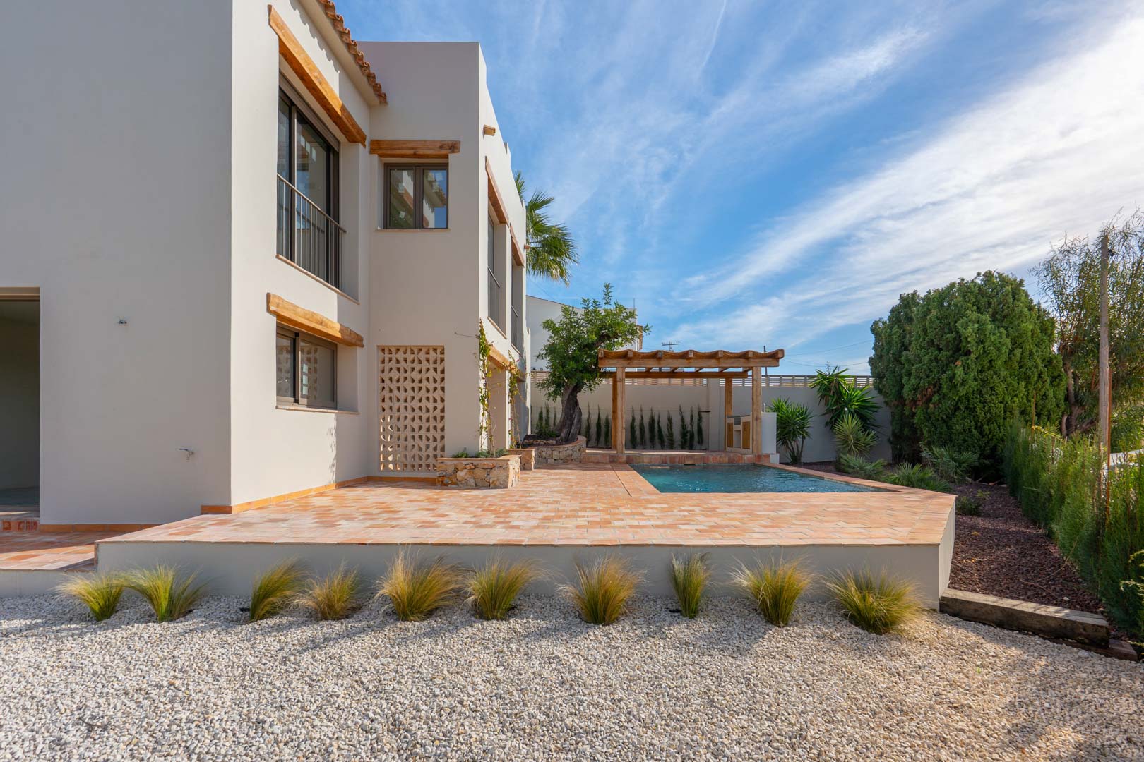 Exterior view featuring a terracotta terrace, a newly built swimming pool, and a wooden pergola set within a landscaped Mediterranean garden with gravel and ornamental grasses.