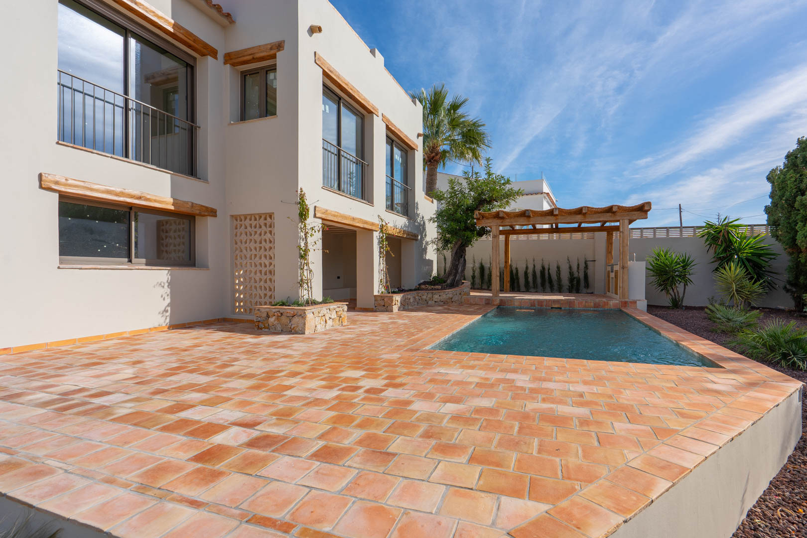 Exterior view featuring a terracotta tile terrace, private swimming pool, and wooden pergola. The facade includes natural wood lintels and integrated stone planters.
