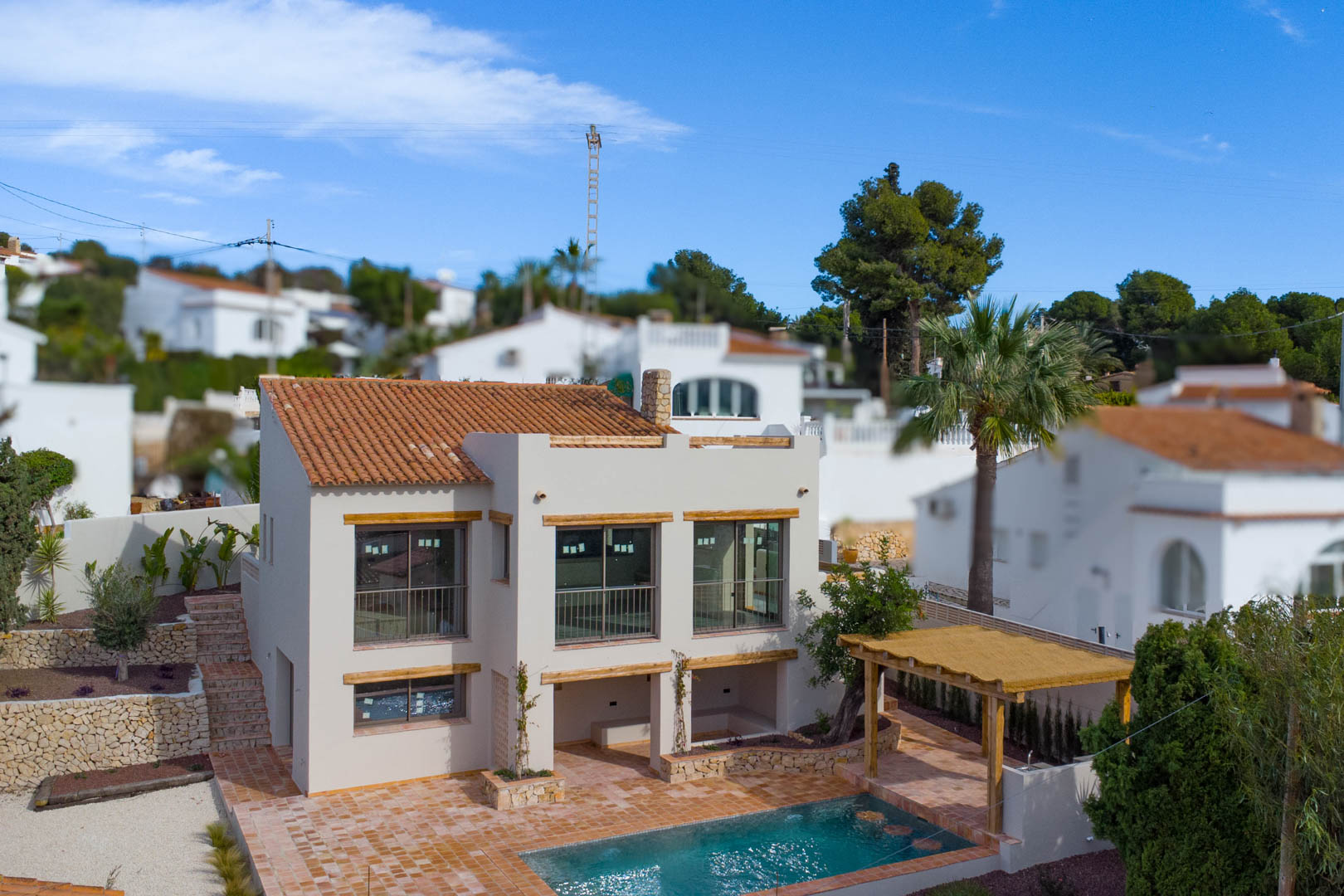 Exterior view of the Mediterranean villa featuring large windows, terracotta roofing, a private pool area with a wooden pergola, and natural stone walls.