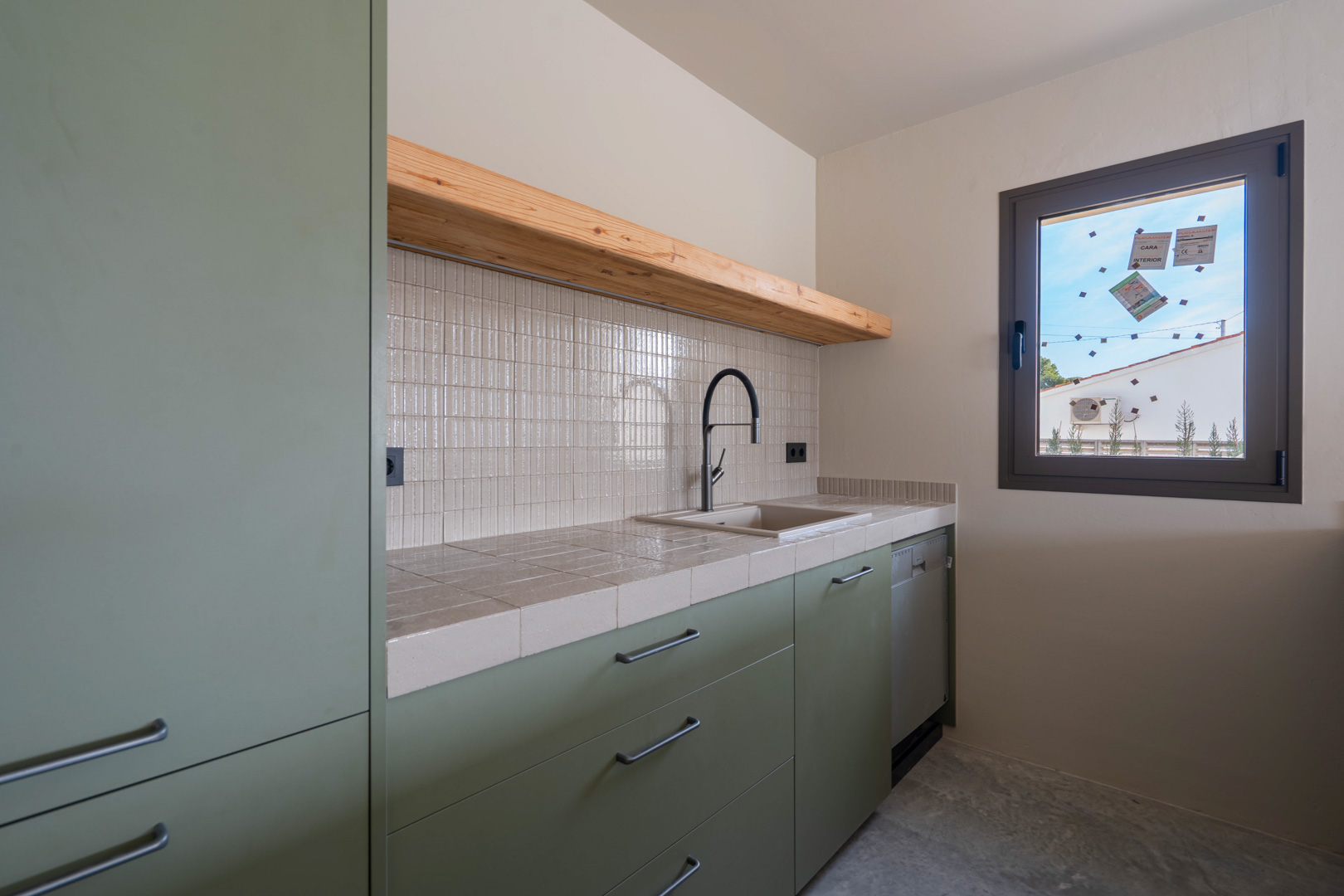 Guest kitchenette featuring sage green cabinetry, a tiled countertop, solid wood shelving, and a black designer faucet against clay-finished walls.