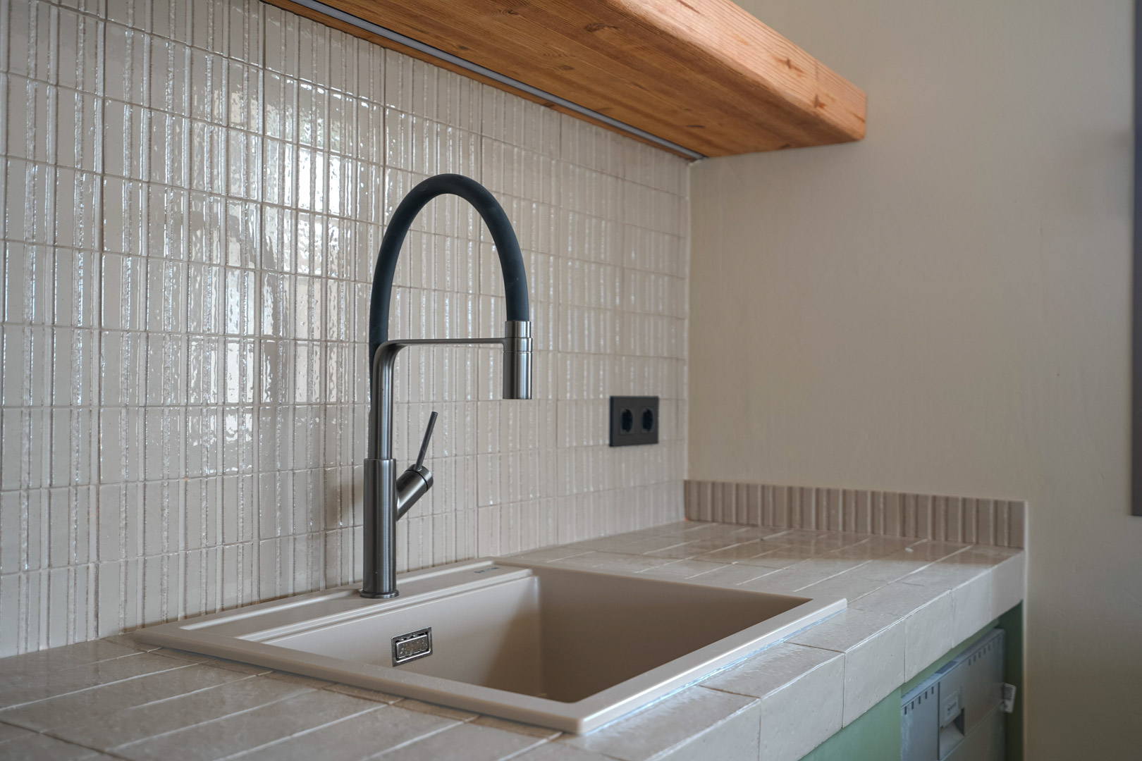 Kitchen detail featuring a porcelain tile countertop with an integrated sink. Modern flexible faucet set against a vertical mosaic backsplash and solid wood shelving.