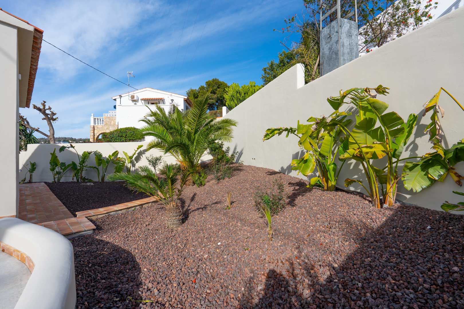 Mediterranean garden area featuring volcanic gravel, palms, and banana plants bordered by a white wall and terracotta tile walkway.