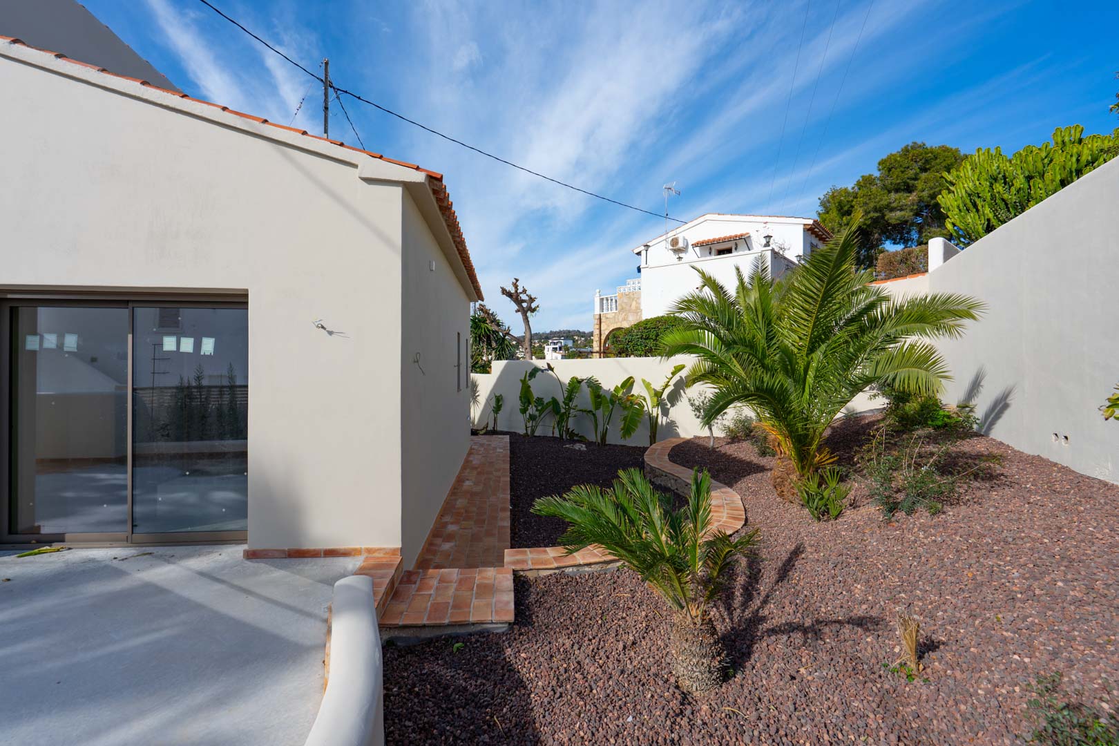 Mediterranean garden featuring palm trees and volcanic gravel, bordered by terracotta tile paths and white perimeter walls.