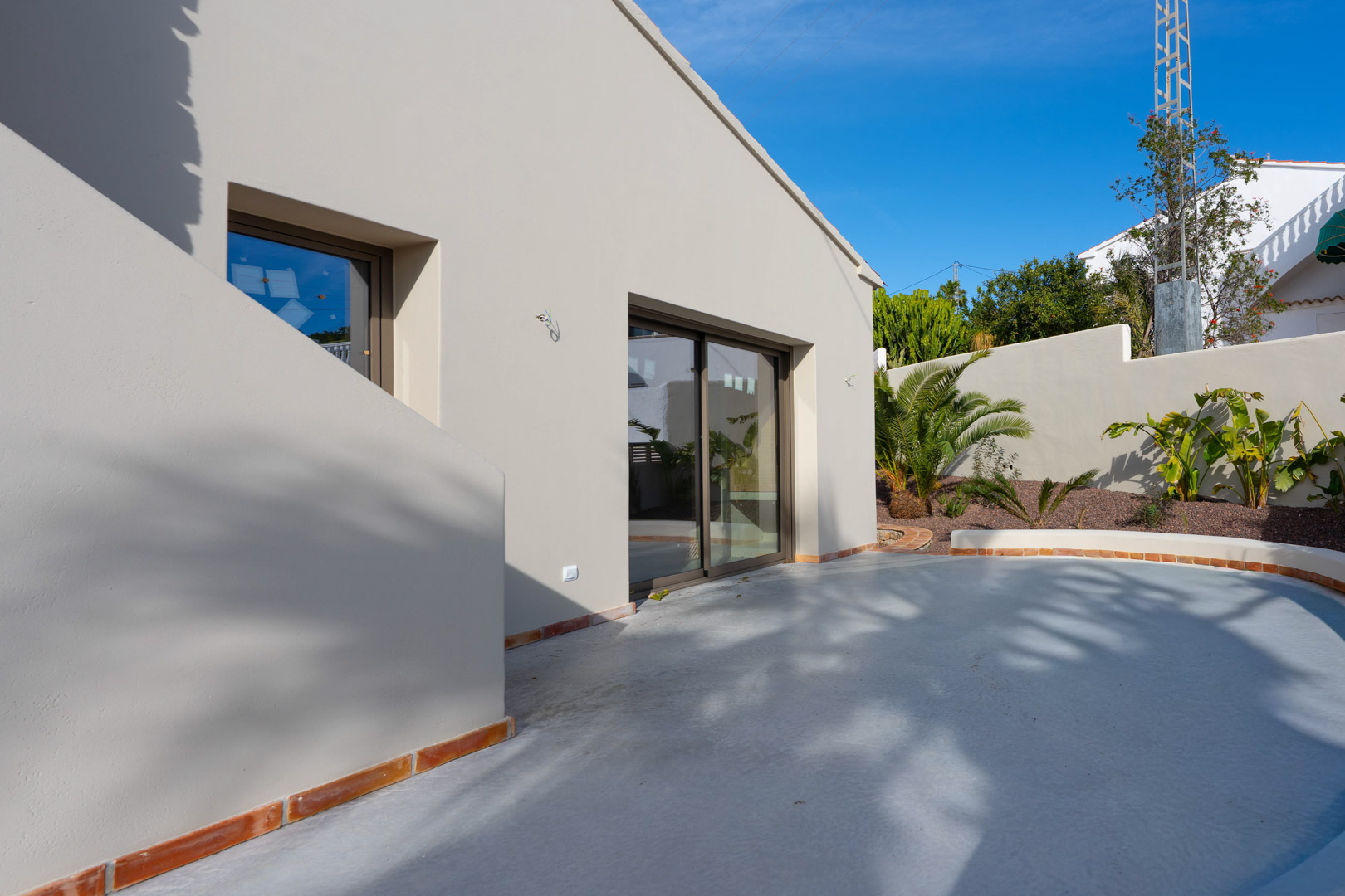 Minimalist facade with earth-toned finishes, large sliding glass doors, and a curved terrace featuring terracotta edging and Mediterranean landscaping.