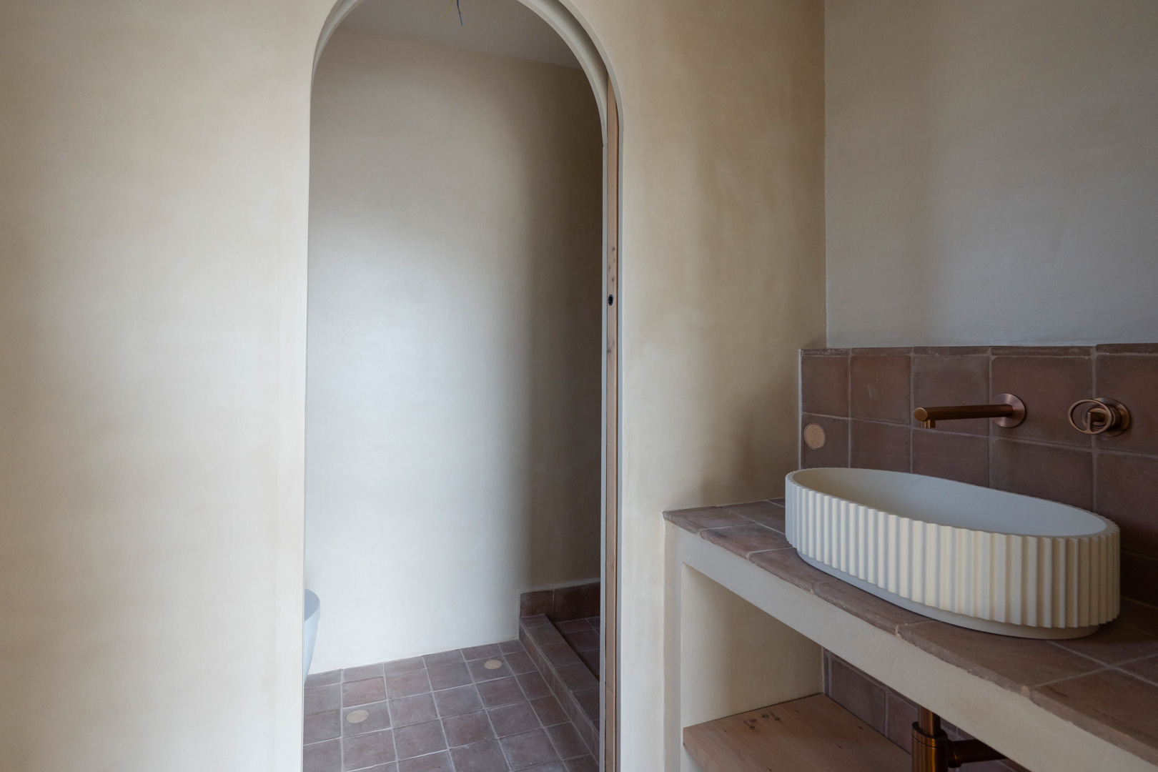 Modern bathroom featuring clay-finished walls, a fluted vessel sink, and bronze wall-mounted fixtures. An elegant archway leads to the shower area with terracotta tiling.