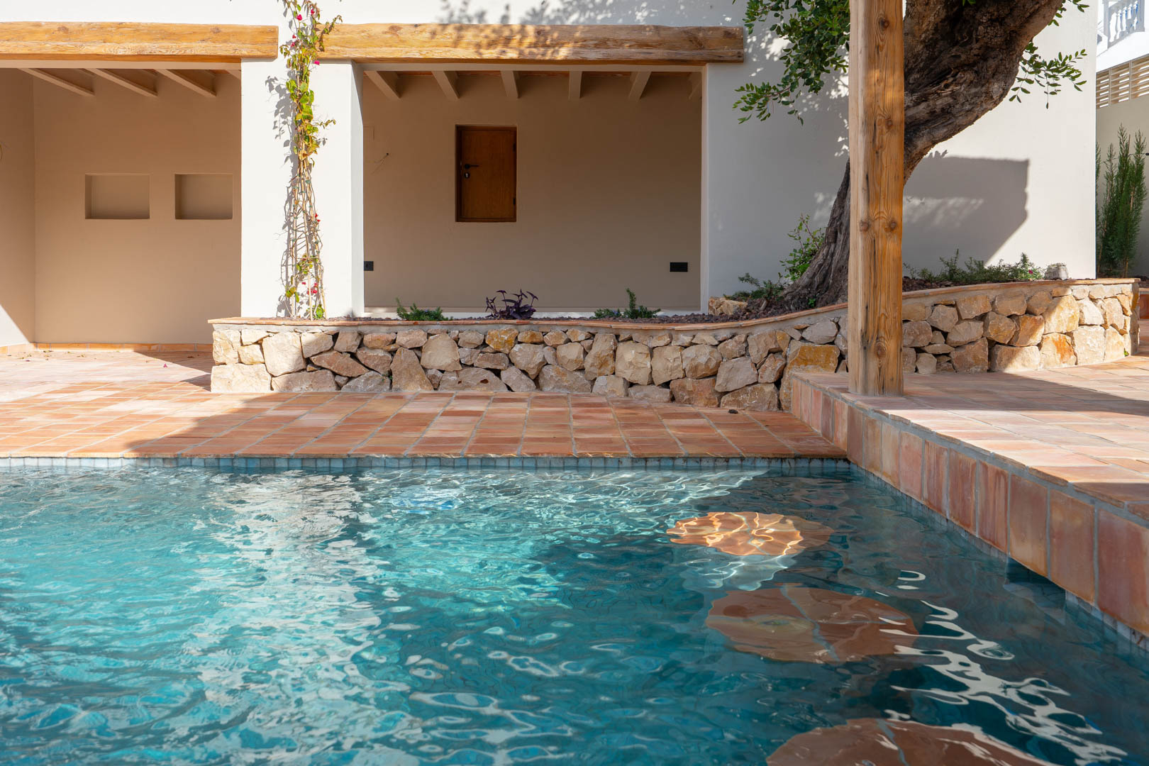 Newly built pool featuring integrated underwater seating, terracotta tiling, natural stone walls, and rustic wooden beams framing the shaded porch area.