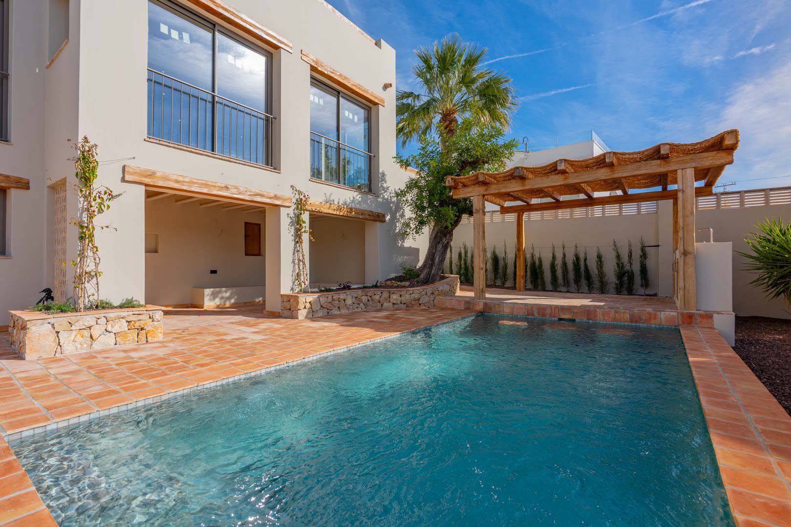 Newly built swimming pool featuring terracotta tiling, a wooden pergola, and natural stone accents integrated into the villa's Mediterranean facade.