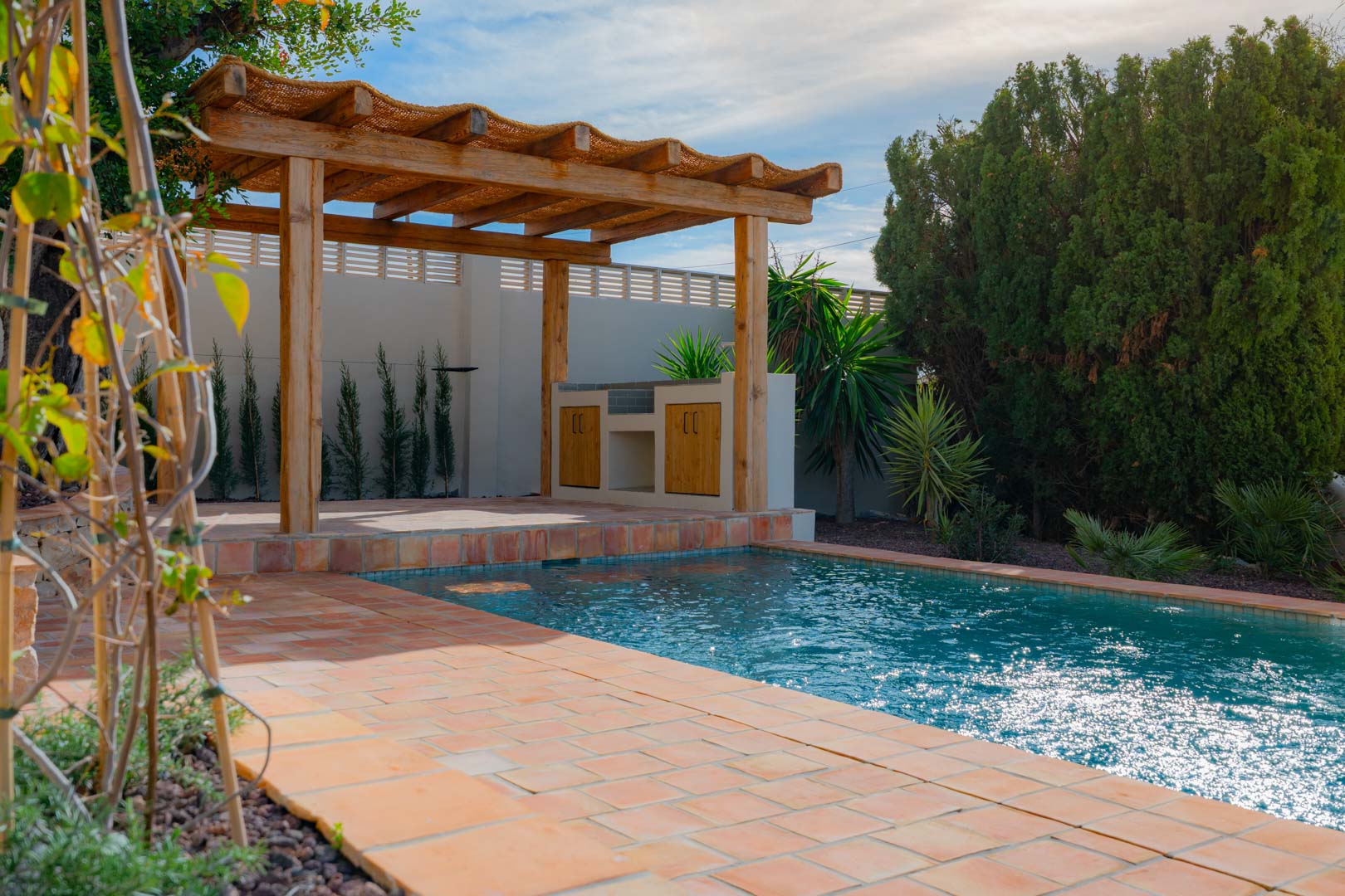 Outdoor area featuring a blue-tiled swimming pool, terracotta paving, and a wooden pergola with an integrated summer kitchen surrounded by Mediterranean landscaping.