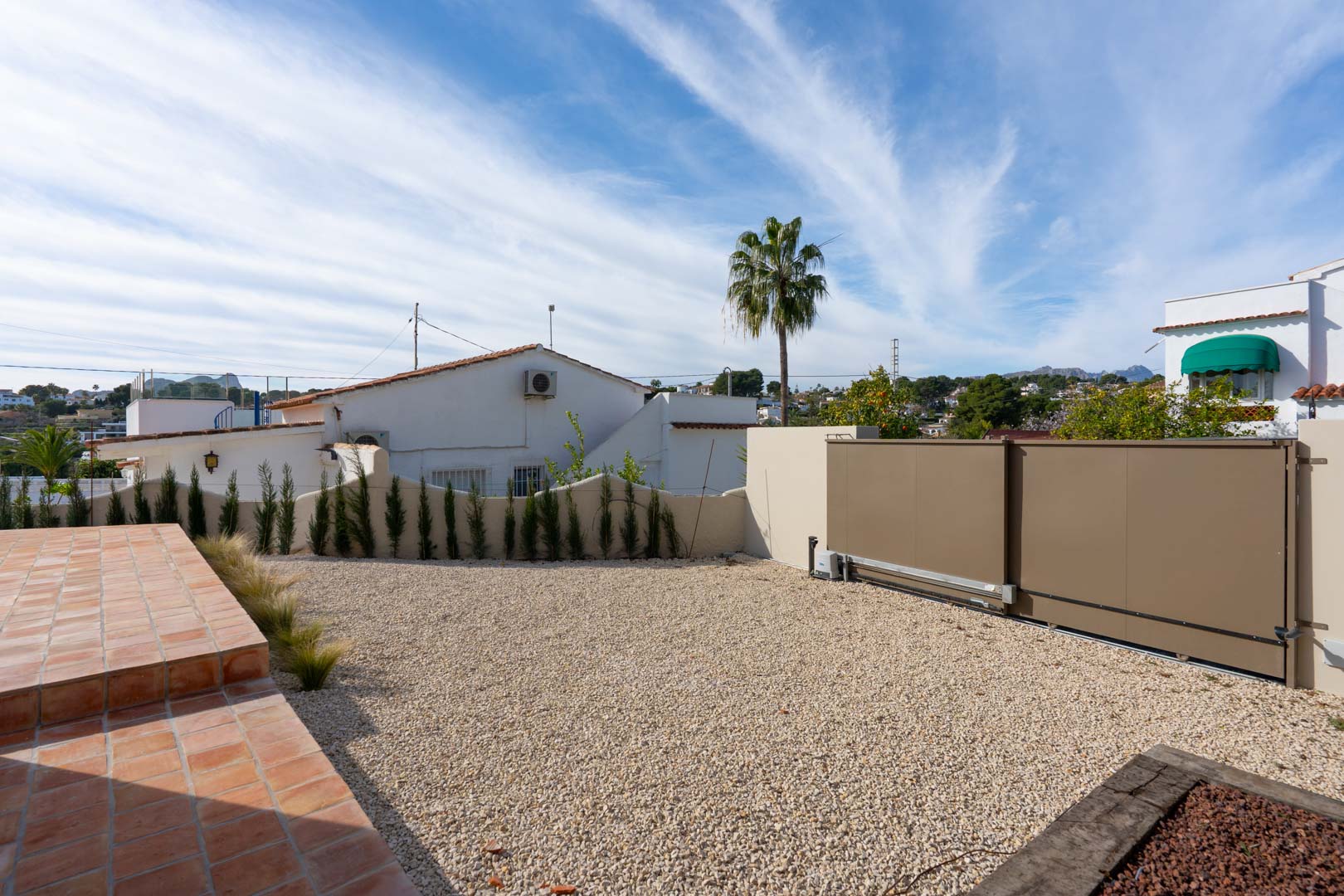 Outdoor parking area featuring a gravel driveway, motorized sliding gate, and terracotta tiled steps, bordered by young cypress trees.