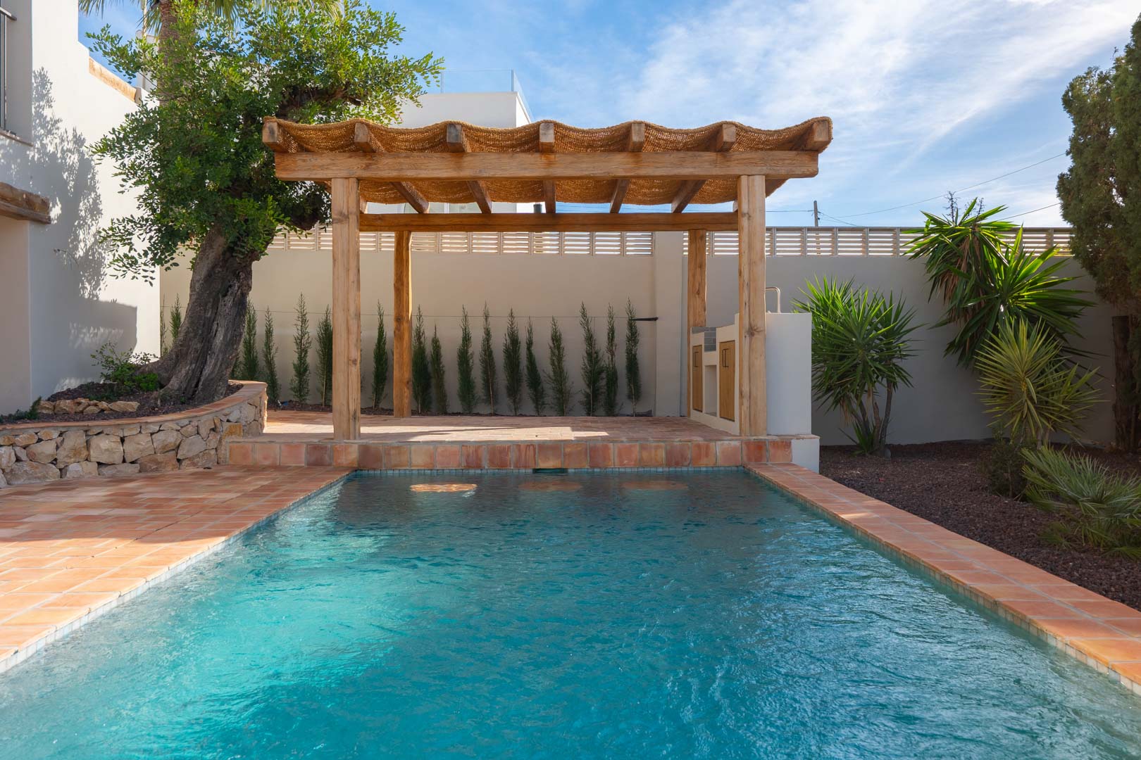 Outdoor swimming pool with terracotta tiling and a rustic wooden pergola featuring an integrated BBQ area, set within a landscaped Mediterranean garden.