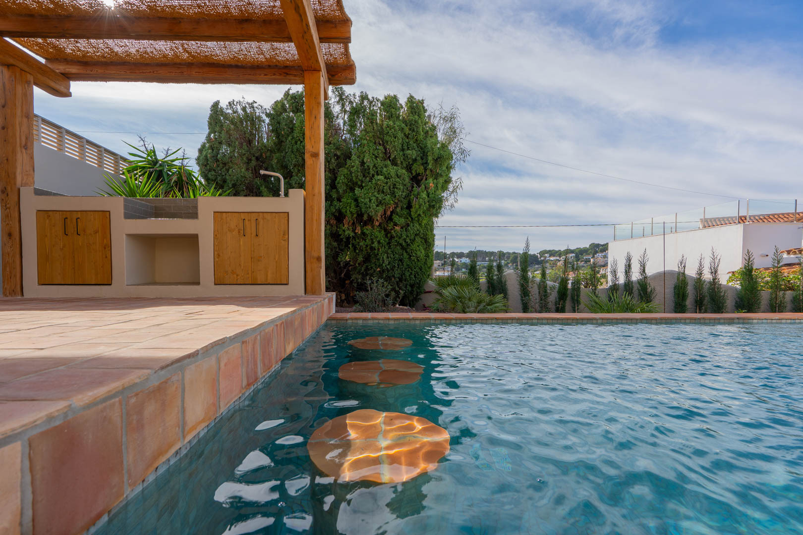 Pool area featuring submerged seating, an outdoor kitchen with wooden cabinetry under a rustic pergola, and traditional terracotta tiling.