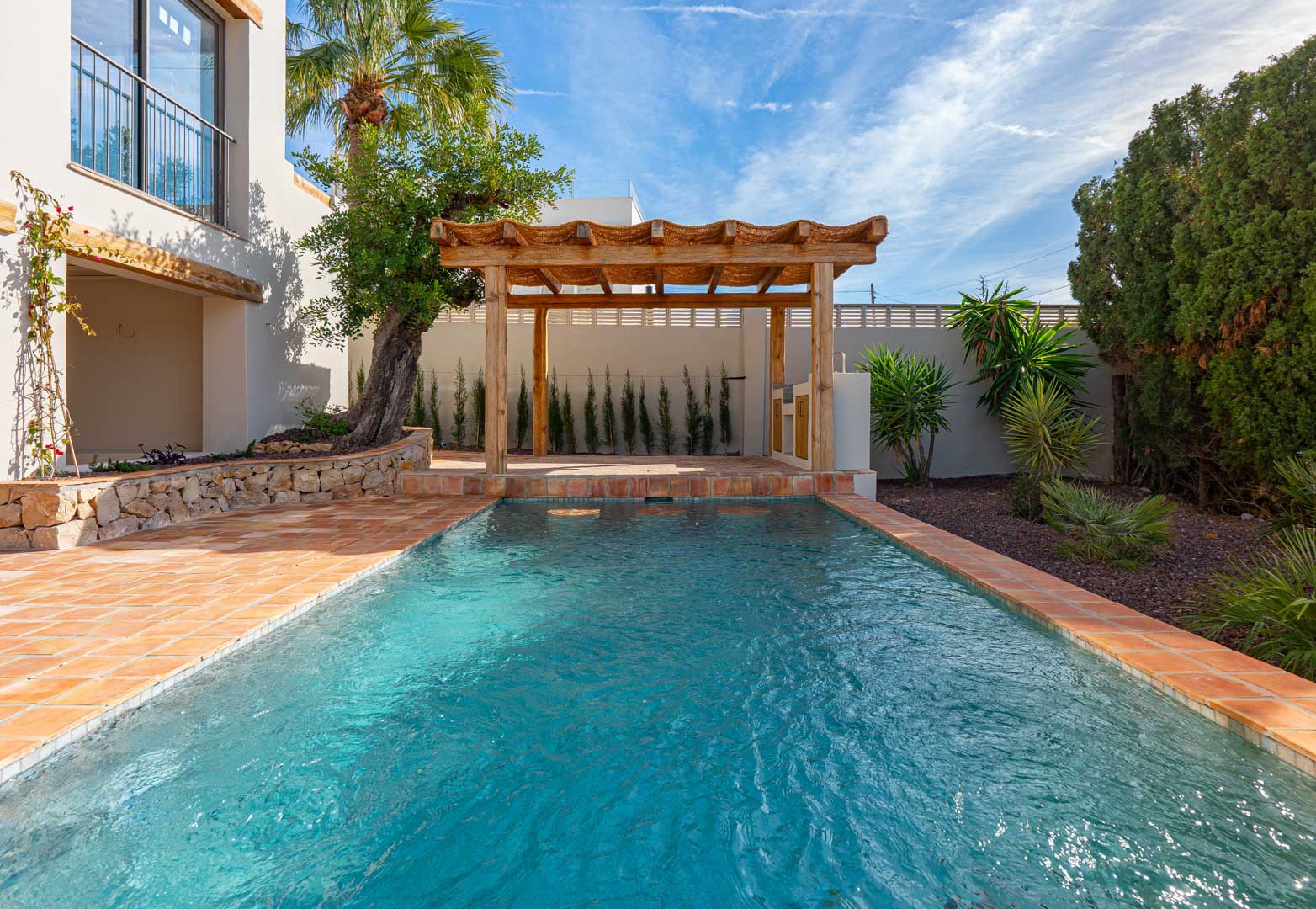 Rectangular swimming pool with terracotta surrounds, a wooden pergola, and a landscaped Mediterranean garden featuring stone walls and native palm trees.