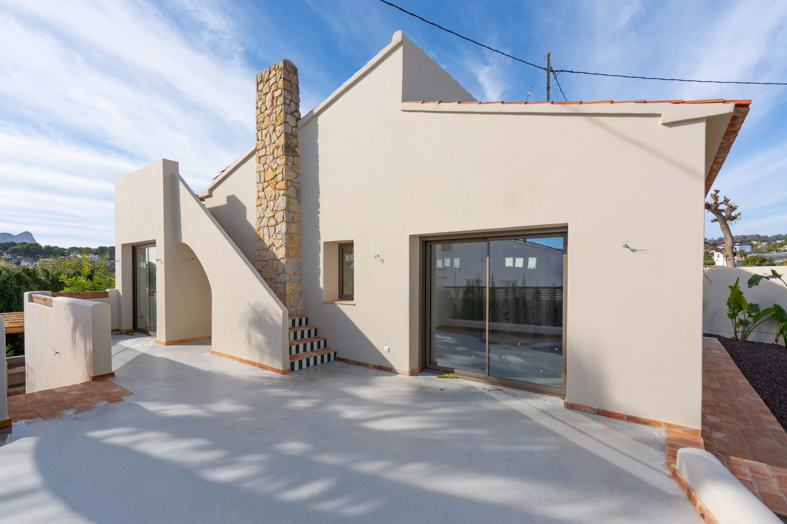 Villa exterior featuring a polished concrete terrace with terracotta borders, a natural stone chimney, and views of the Peñón de Ifach in the distance.