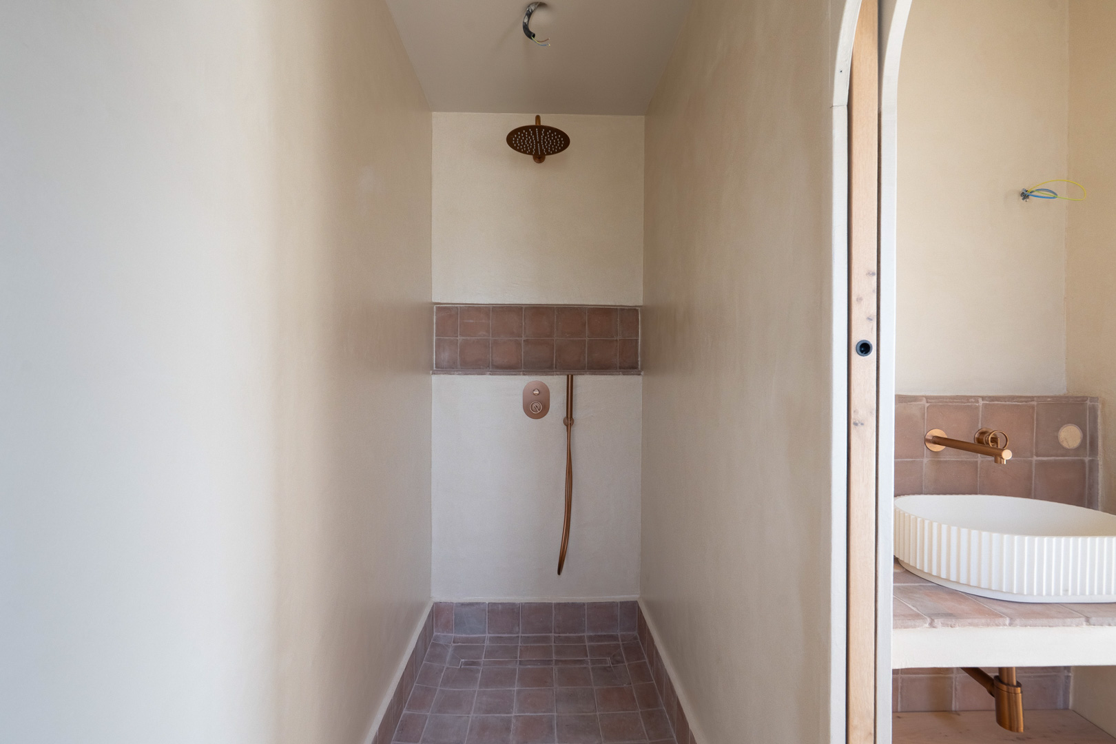 Walk-in shower featuring natural clay wall finishes, bronze-toned fixtures, and terracotta tiling. Adjacent vanity shows a fluted vessel sink with wall-mounted taps.