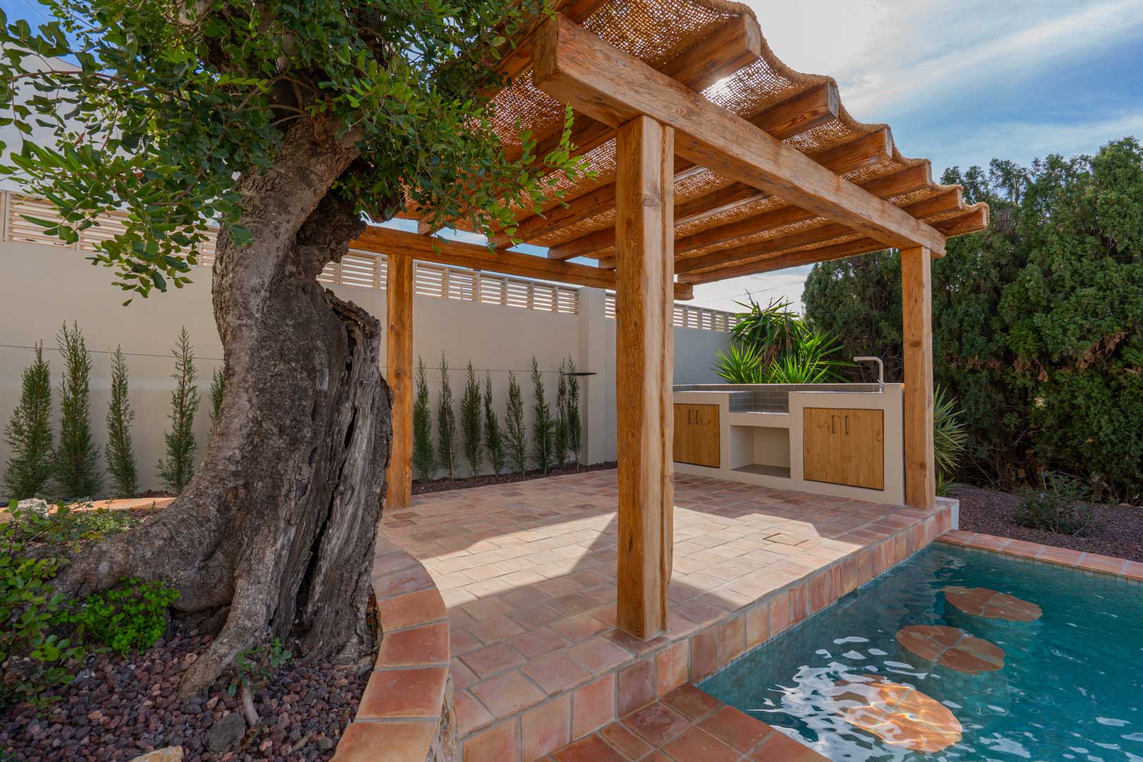 Wooden pergola with a natural fiber roof on terracotta tiles, featuring an outdoor kitchen and a pool with submerged seating next to a mature olive tree.