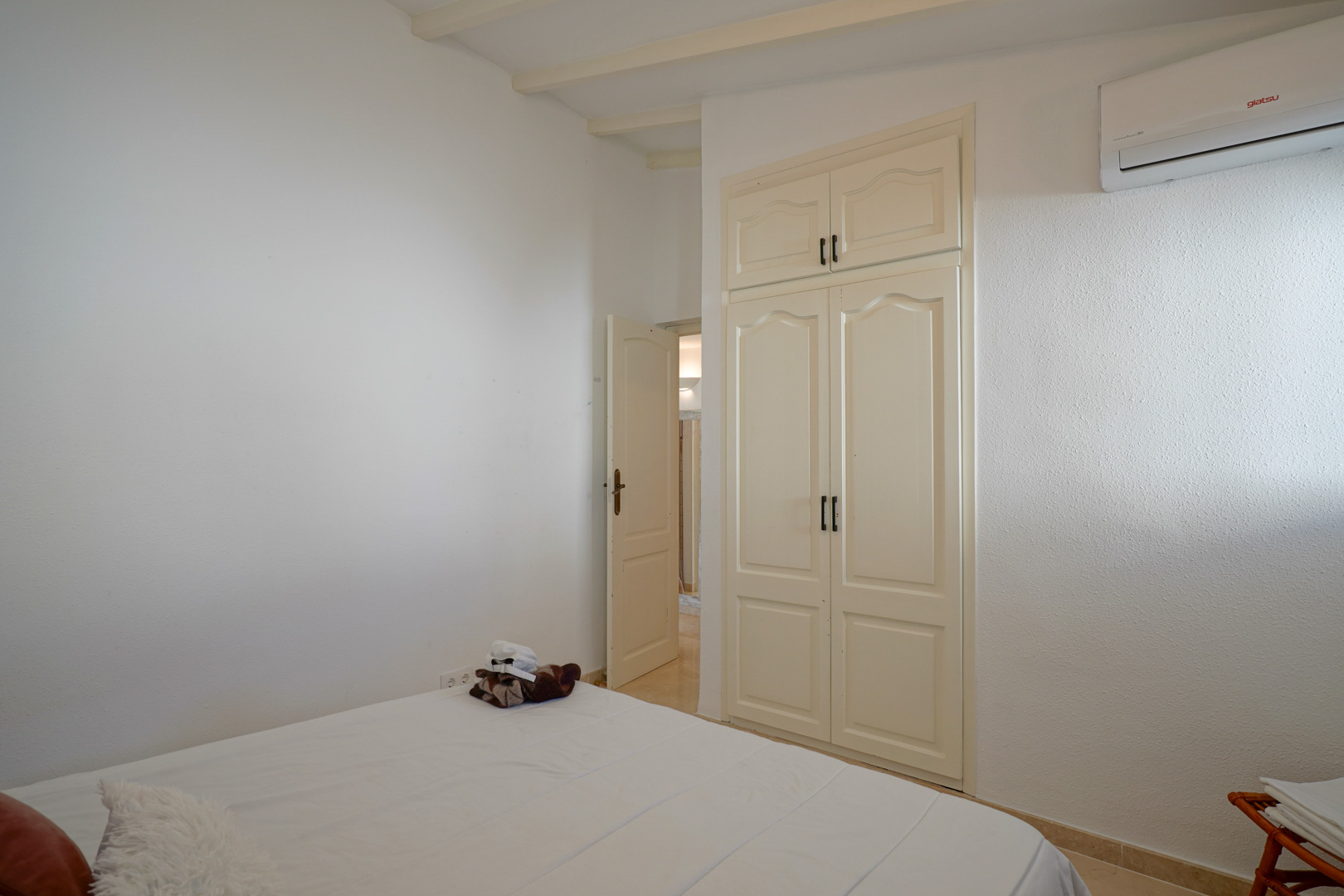 Bright bedroom featuring a built-in wardrobe with traditional doors, exposed ceiling beams, and a wall-mounted air conditioning unit.