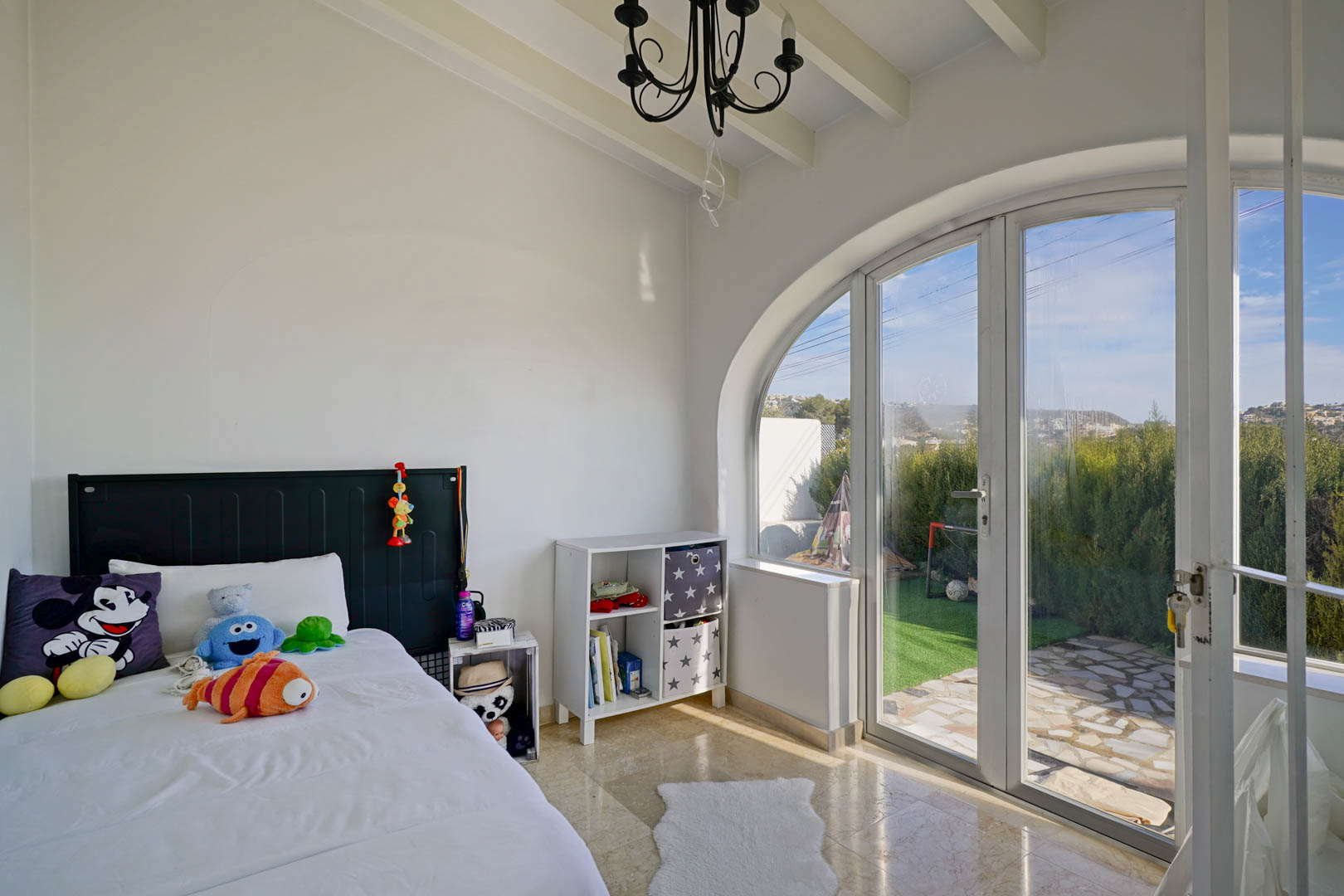 Bright bedroom featuring white-beamed ceilings and a large arched glass door providing direct access to the garden and ample natural light.