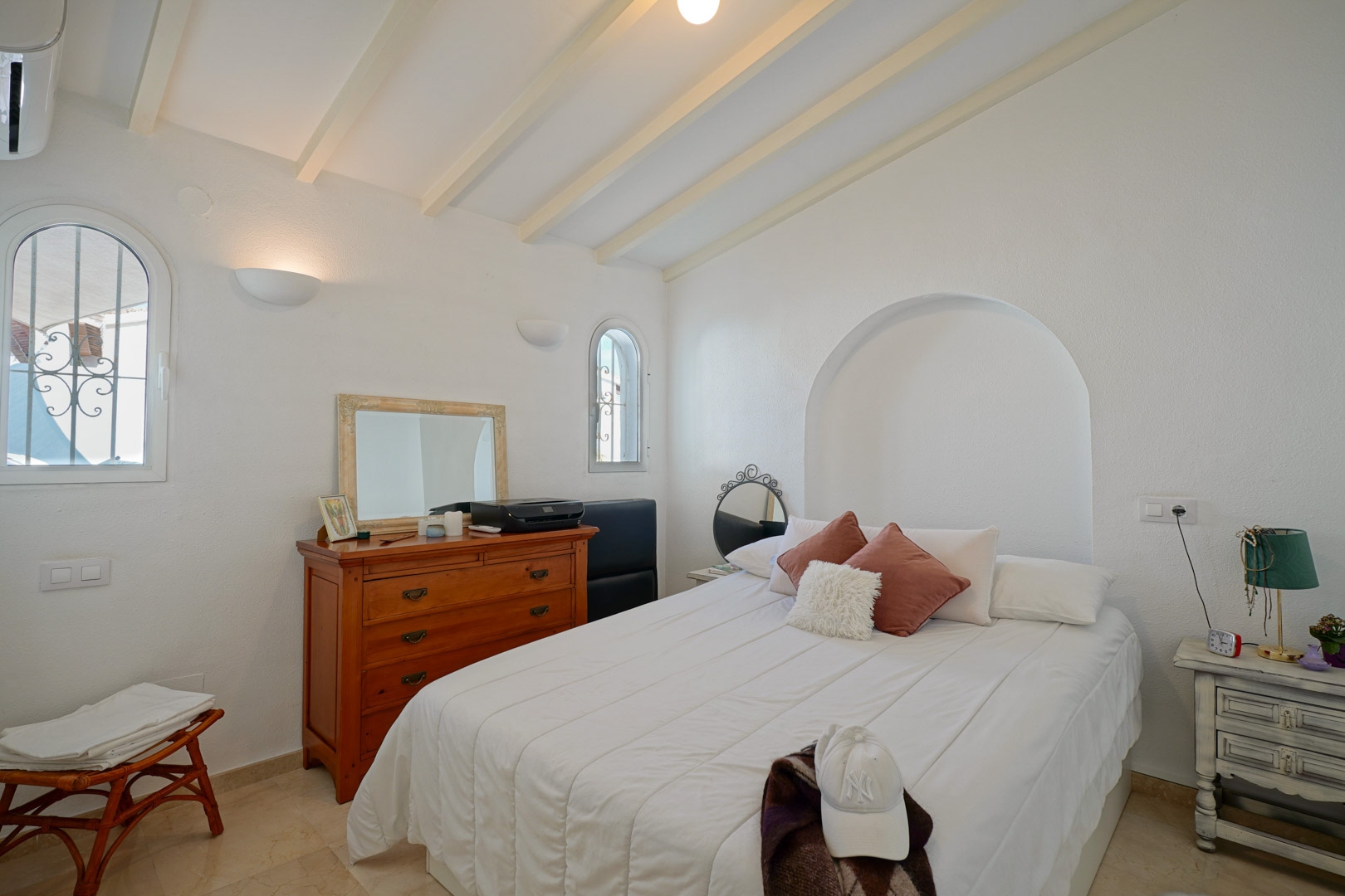 Bright bedroom featuring white-painted ceiling beams, arched windows with security grills, and a decorative wall niche. Equipped with air conditioning and marble floors.