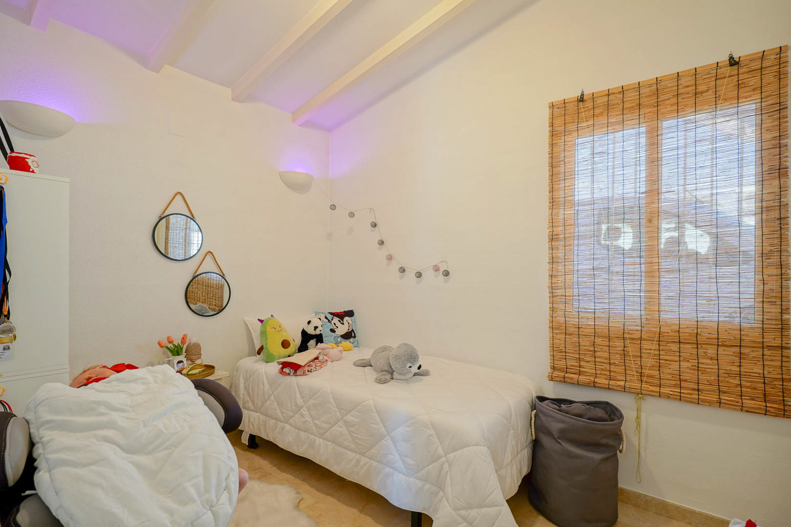 Cozy bedroom featuring white-painted ceiling beams, a single bed, decorative rope-hung mirrors, and a natural fiber blind for a warm atmosphere.