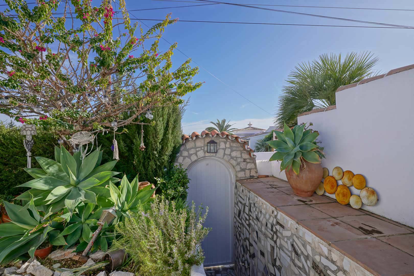 Exterior entrance featuring a natural stone archway, light grey gate, and Mediterranean flora including agaves and bougainvillea against white walls.