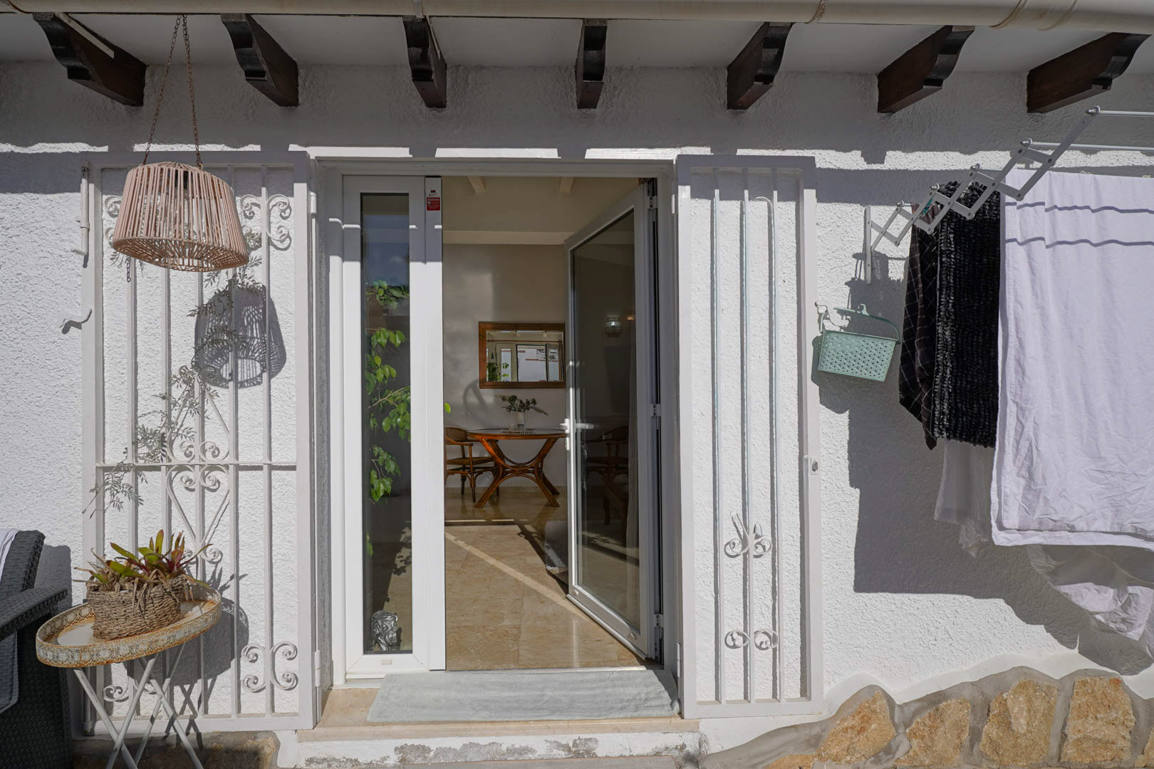 Exterior entrance featuring a white glass door with security grilles under traditional wooden beams. The view reveals a glimpse of the interior dining area and polished tiled flooring.