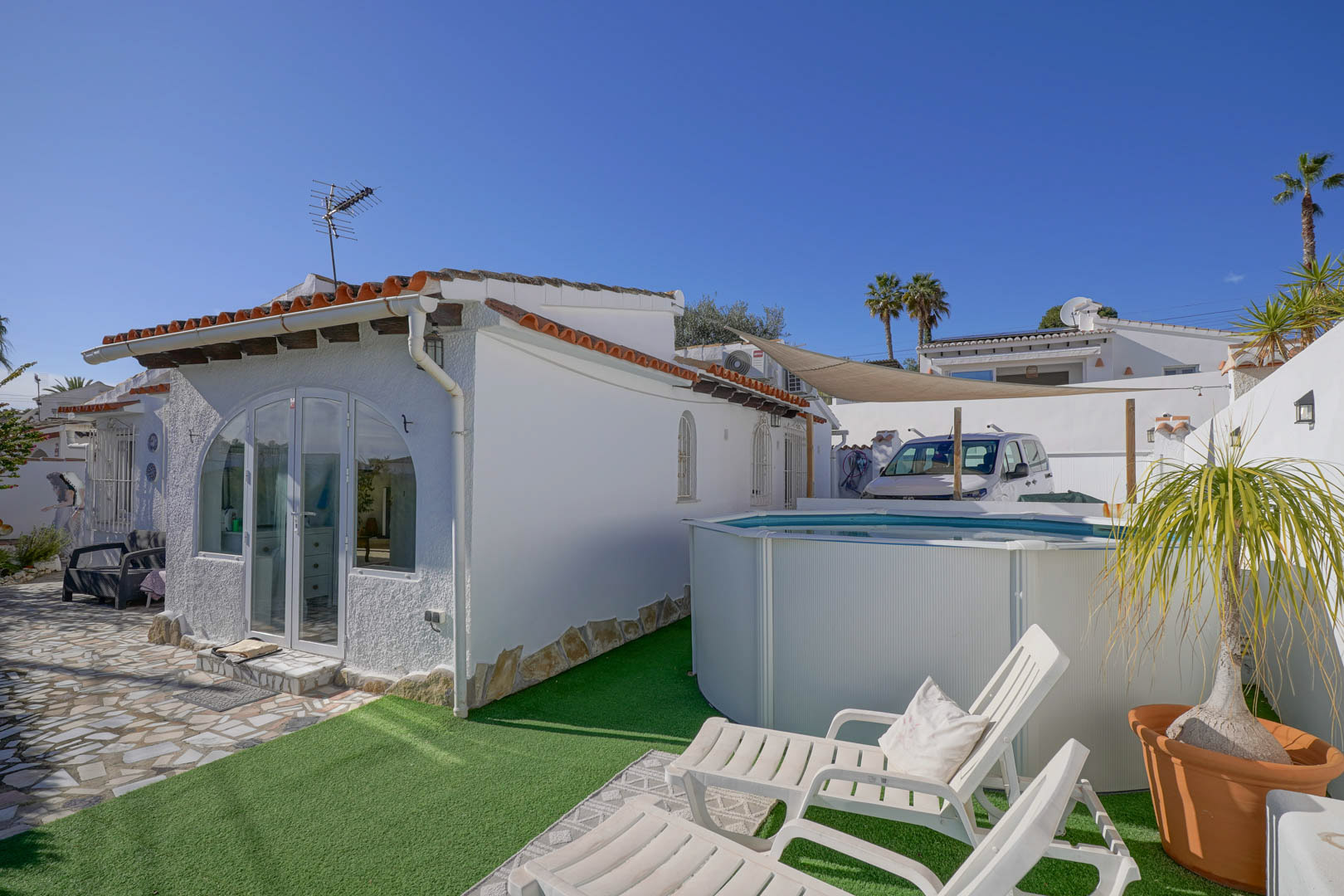 Exterior view of the white villa featuring a glazed porch, a portable swimming pool on artificial turf, and a sunbathing area with white loungers.