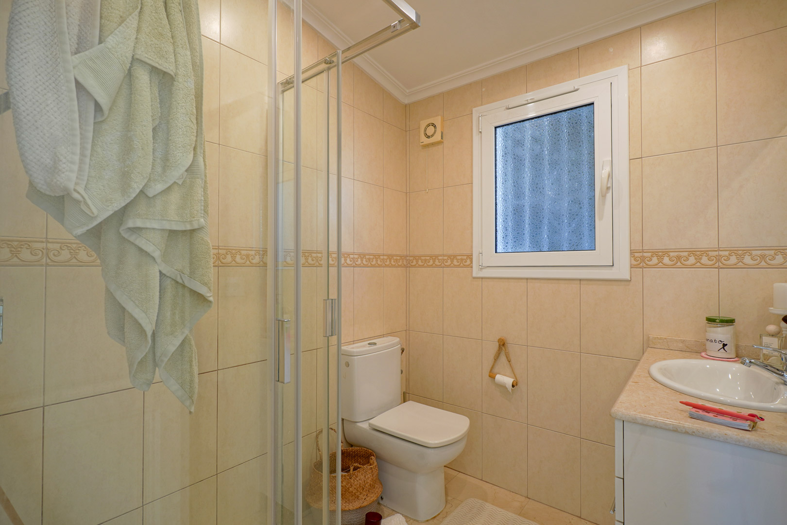 Full bathroom featuring cream-toned tiled walls, a glass shower enclosure, white ceramic fixtures, and a vanity with a marble-effect countertop.