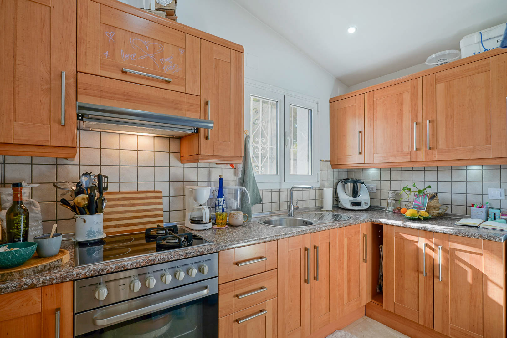 Independent kitchen featuring wood-finish cabinetry, granite countertops, and a hybrid hob. Includes a circular stainless steel sink and a window providing natural light.
