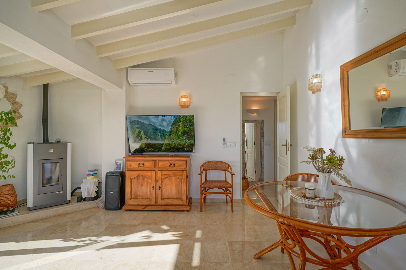 Living-dining area featuring white exposed ceiling beams, marble floors, a pellet stove, and rattan furniture. The space includes air conditioning and receives ample natural light.