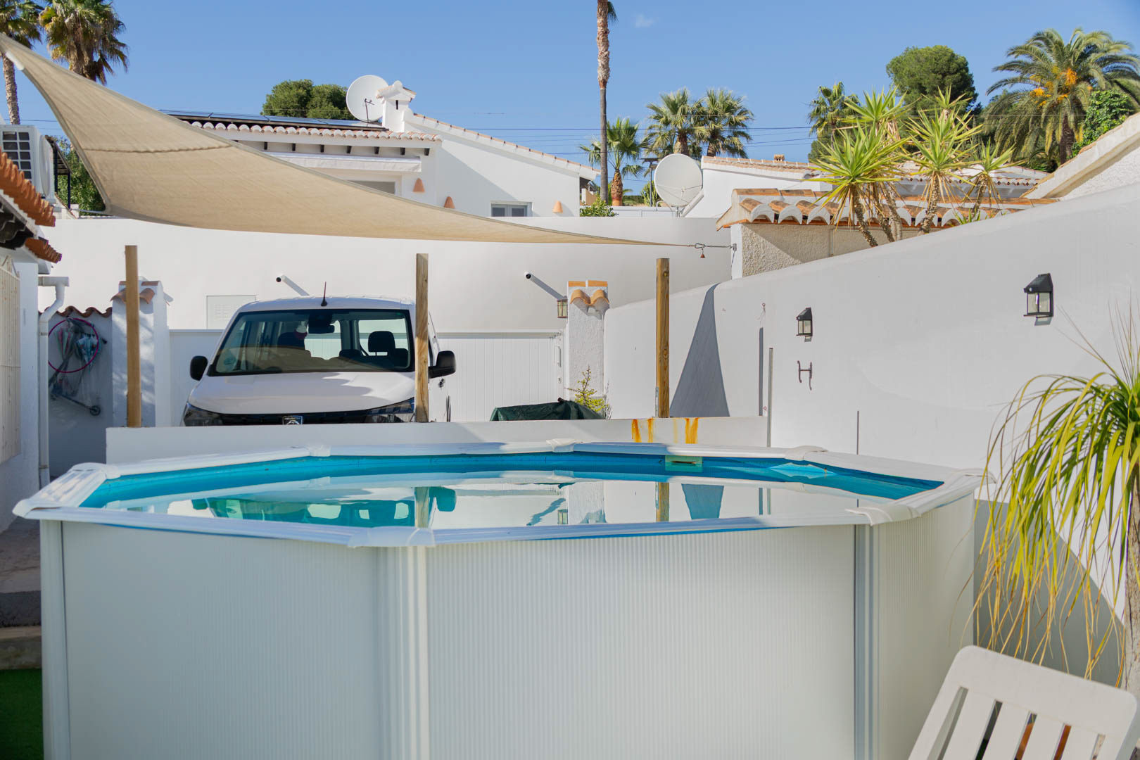 Octagonal portable pool on a sunny terrace featuring a beige sun sail, surrounded by white privacy walls and Mediterranean palm trees.