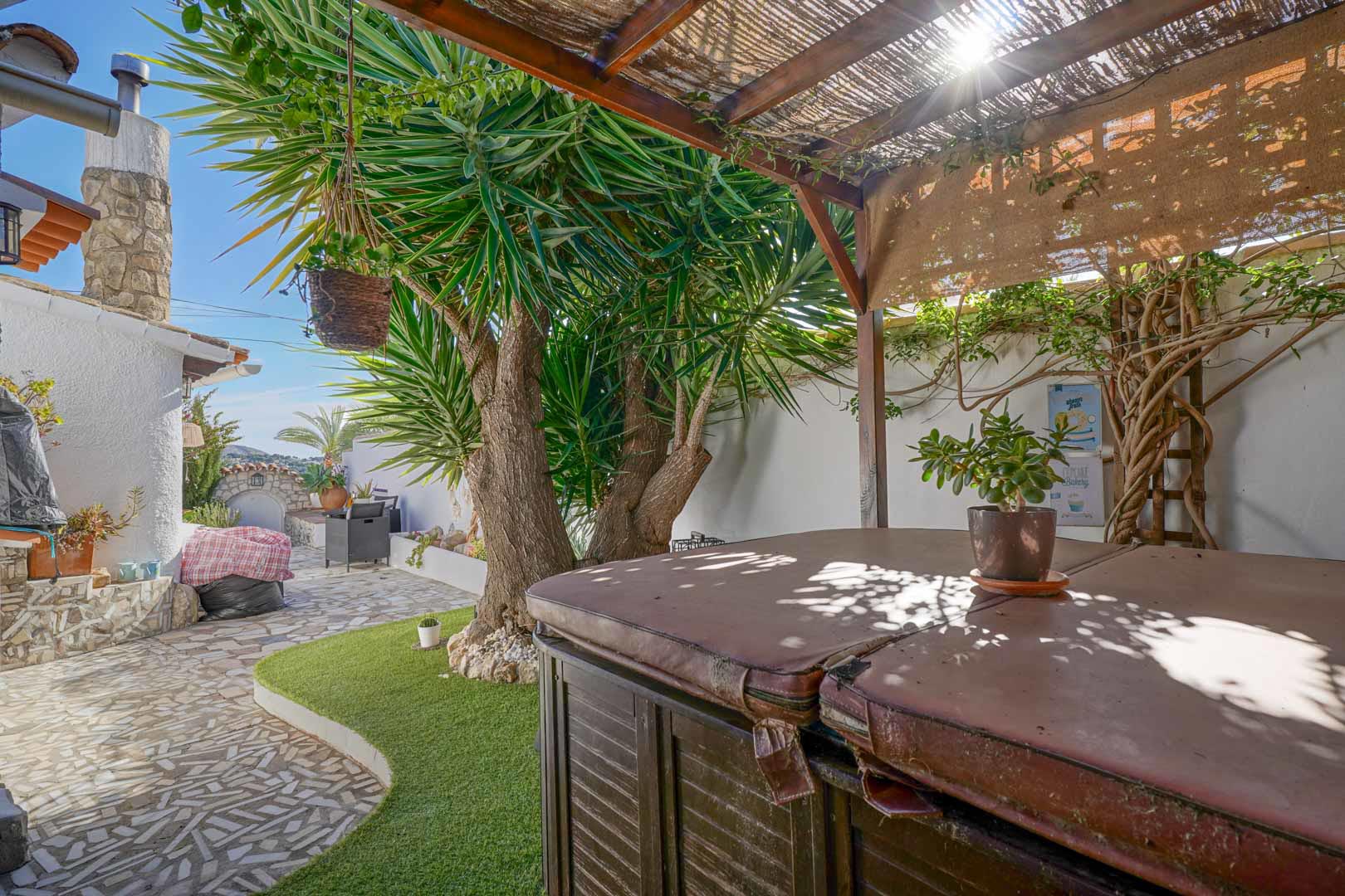 Outdoor terrace featuring a covered jacuzzi under a wooden pergola, set within a low-maintenance garden with stone paving and mature Mediterranean plants.