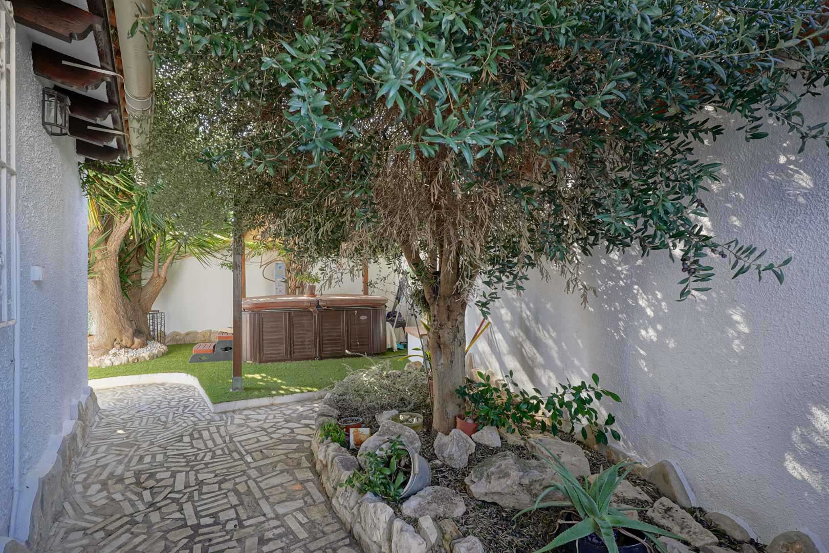 Outdoor terrace featuring a private hot tub on artificial turf, framed by mature Mediterranean trees and a natural stone-paved pathway.