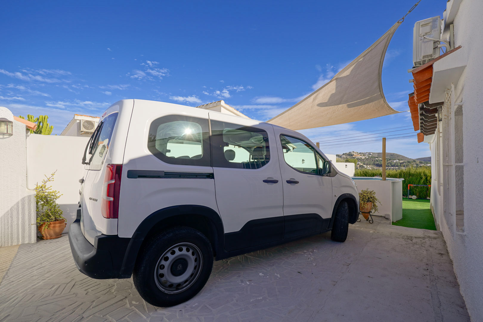 Private parking area featuring patterned paving and a shade sail for sun protection, located alongside the villa's white exterior with surrounding hill views.