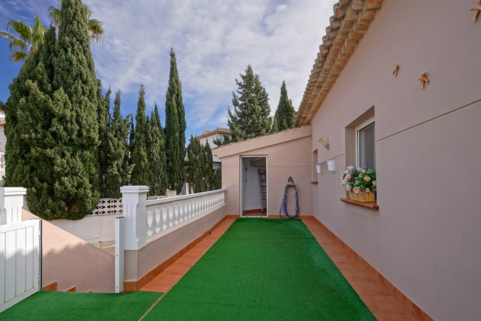 Access terrace featuring artificial turf, white balustrades, and terracotta tiling, leading to the laundry room amidst Mediterranean greenery.