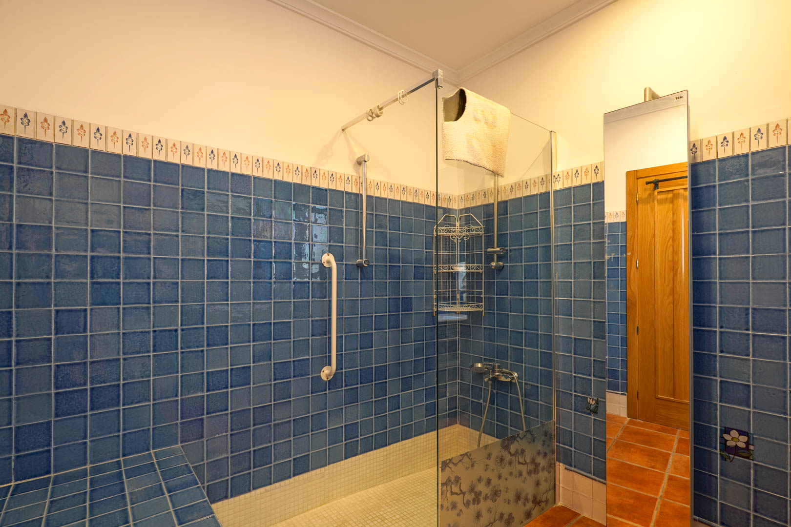 Bathroom featuring a walk-in shower with blue tiling, glass partition with floral accents, white mosaic floor, and a safety handrail for accessibility.