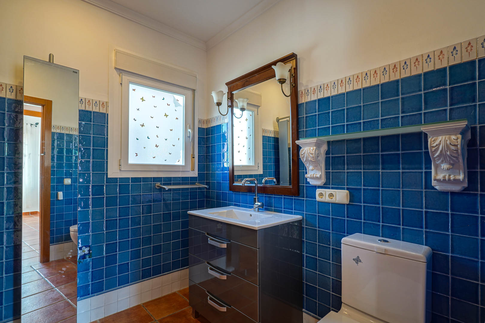 Bathroom featuring vibrant blue wall tiles with decorative borders, a modern dark vanity unit, and traditional terracotta flooring under natural light.