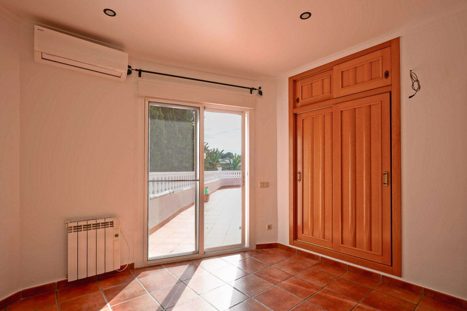 Bedroom featuring a built-in wooden wardrobe, terracotta tiling, and terrace access. Equipped with a split AC unit, electric radiator, and recessed ceiling lights.