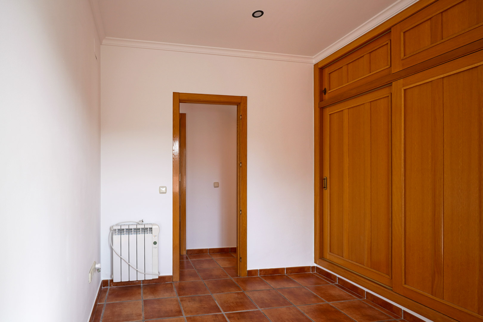 Bedroom featuring a large built-in wooden wardrobe, rustic terracotta floor tiles, and an electric radiator. The space includes white walls and elegant crown molding.