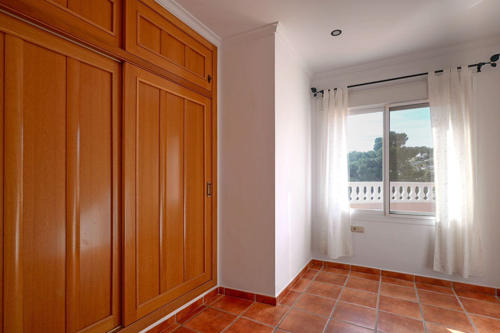 Bedroom featuring a large built-in wooden wardrobe, terracotta tile flooring, and a window overlooking the terrace and Mediterranean landscape.
