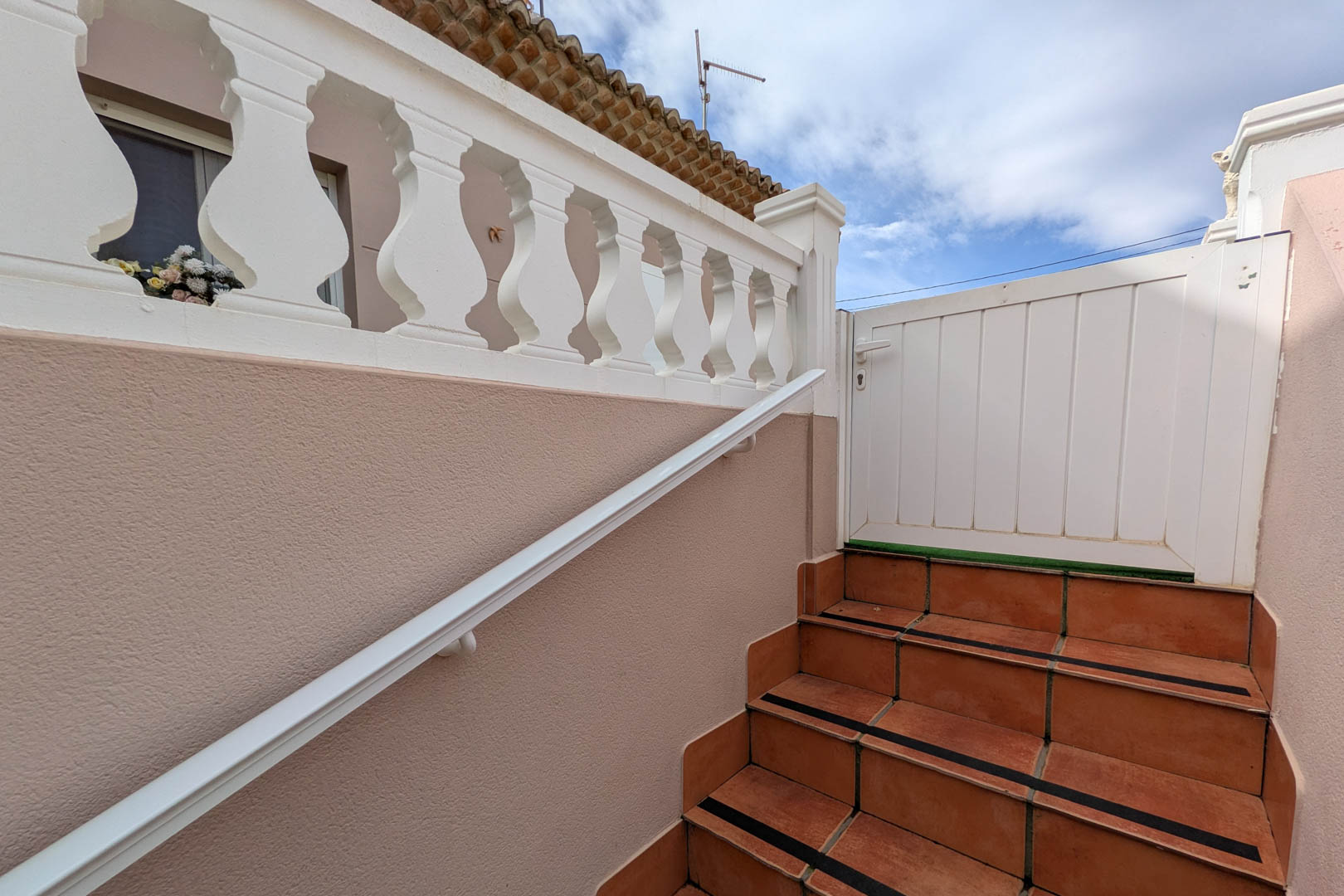 Exterior access staircase featuring terracotta-style tiles with safety strips, a white handrail, and a classic balustrade leading to the upper entrance gate.