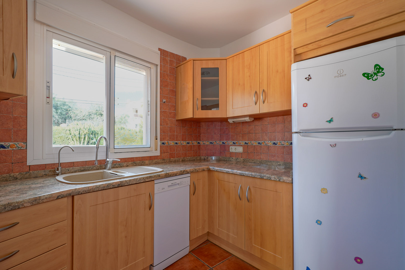 Independent kitchen featuring light wood cabinetry, granite countertops, and rustic tiling. Equipped with a dishwasher, white refrigerator, and a large window for natural light.