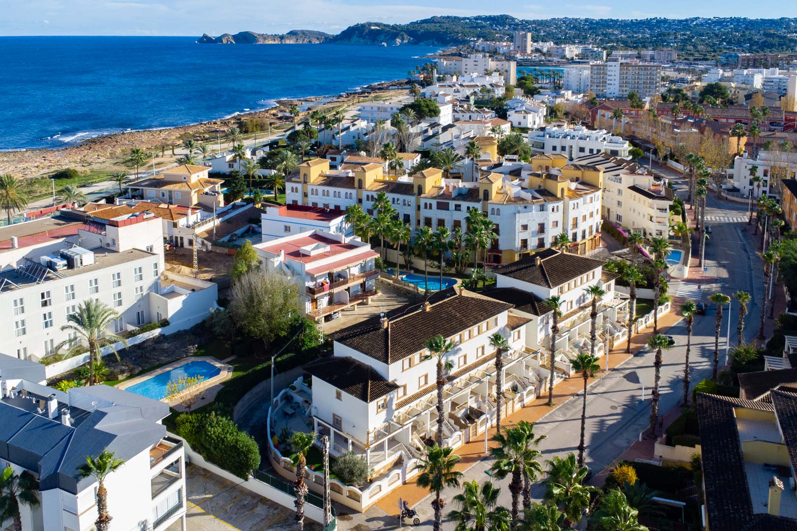 Aerial view of the Calle Génova complex, showing its proximity to the Mediterranean coast, the communal pool, and the white-facade architecture lined with palm trees.